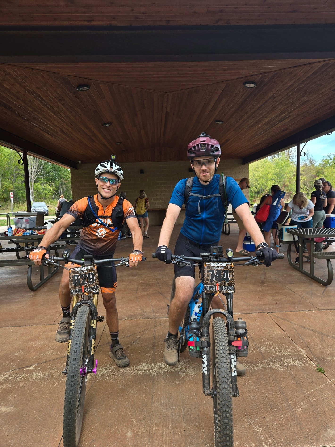 Two mountain bikers on a wooden platform with a wooden roof, surrounded by trees.